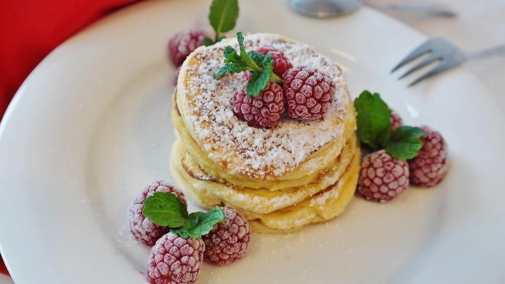 A white plate holds fluffy pancakes topped with fresh raspberries and a dusting of powdered sugar.