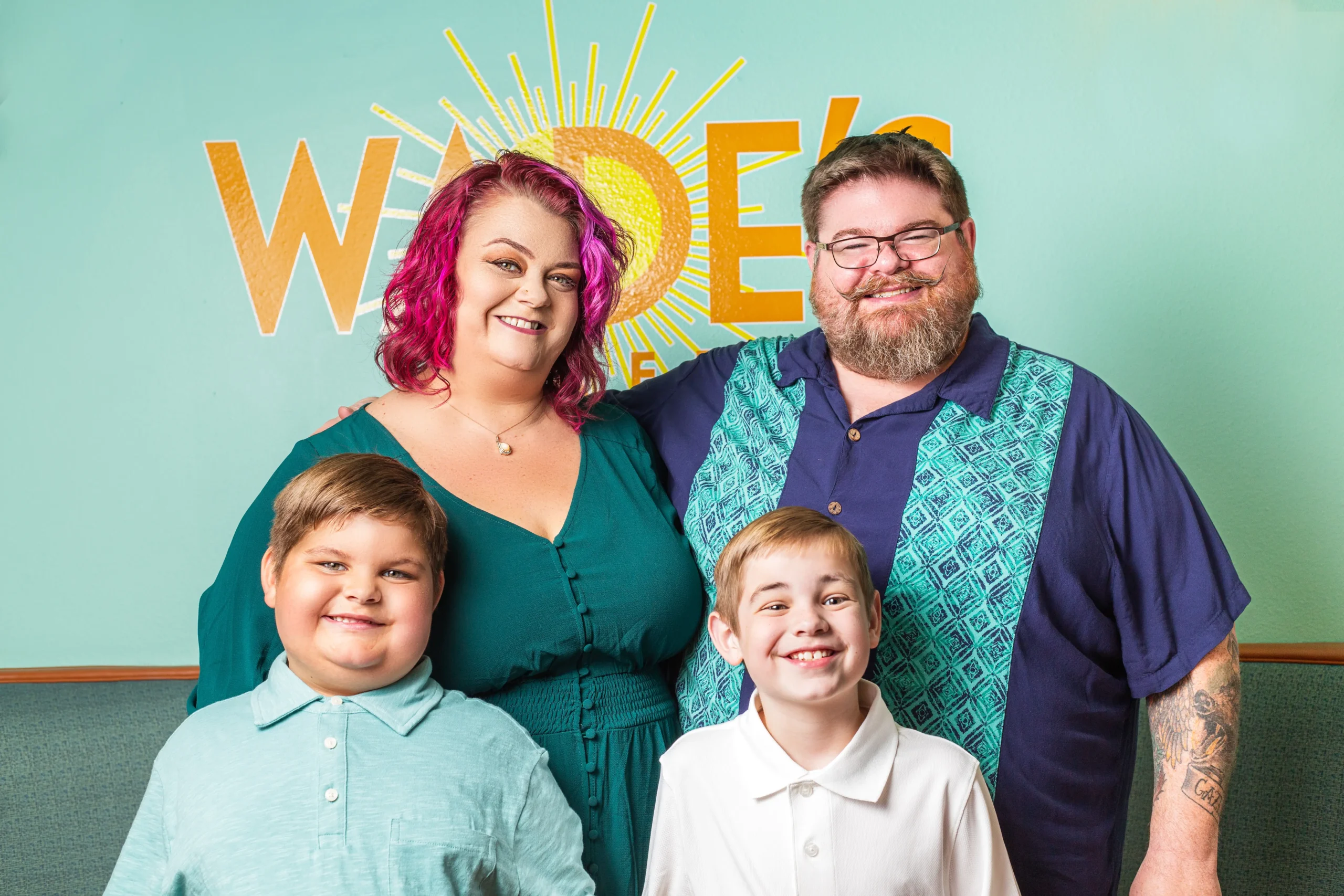 A family stands together smiling for a photo in front of a colorful wall, capturing a moment of joy and togetherness.